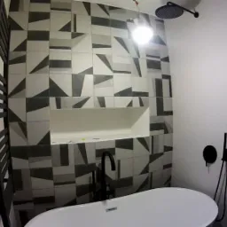 Nowoczesna łazienka z białą wanną wolnostojącą i czarną armaturą na tle ściany wyłożonej płytkami w geometryczne wzory, z wnęką na kosmetyki.