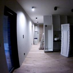 Janos - Wnętrze w trakcie prac wykończeniowych: surowe ściany w odcieniach szarości, drewniana podłoga, widoczne drabiny, zabudowa kuchenna w stanie montażu, drzwi w folii ochronnej.