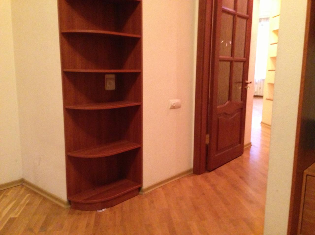 Wnętrze pokoju z narożną, drewnianą półką, brązowymi drzwiami z szybkami i laminowaną podłogą w odcieniach brązu.