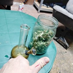 Łukasz Zdeb - Brudny bongo i słoik z suszem na zielonym stole, widoczna ręka z zadrapaniem na kciuku, w tle puszki po piwie i regał.