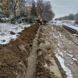 Łukasz Zdeb - Długi, wąski wykop w ziemi pokrytej śniegiem, z pracownikiem w pomarańczowej kamizelce i ciężkim sprzętem w oddali.