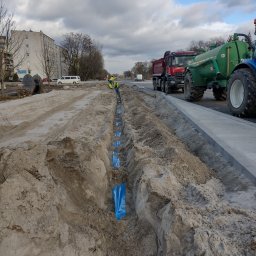 Łukasz Zdeb - Wykopany rów w piasku z niebieskimi osłonami kabli, pracownicy w odblaskowych kamizelkach, ciężarówka i traktor w tle, miejski krajobraz.