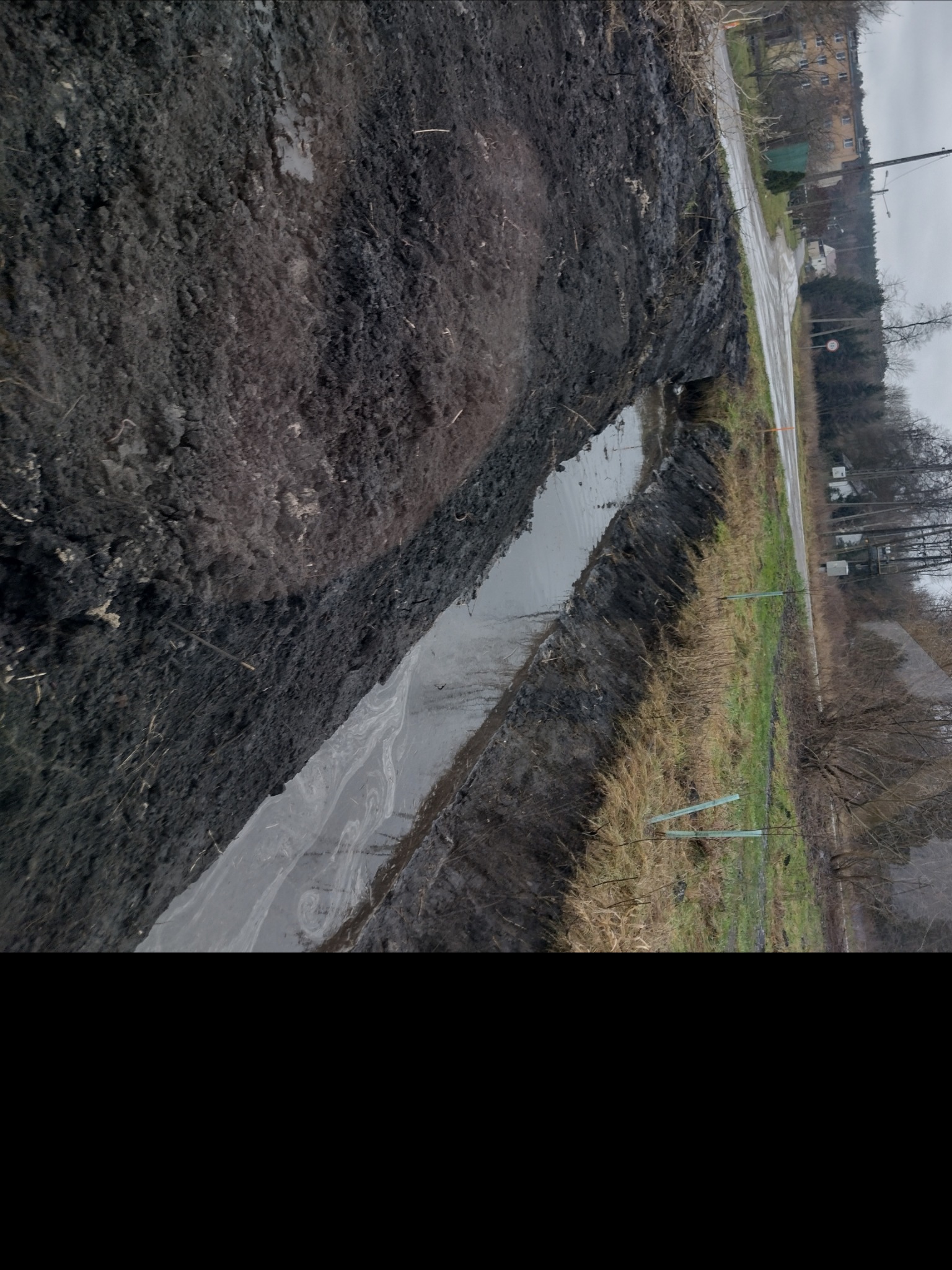 Świeżo wykopany rów melioracyjny wypełniony wodą, widoczna czarna ziemia po bokach, w tle droga i budynki.