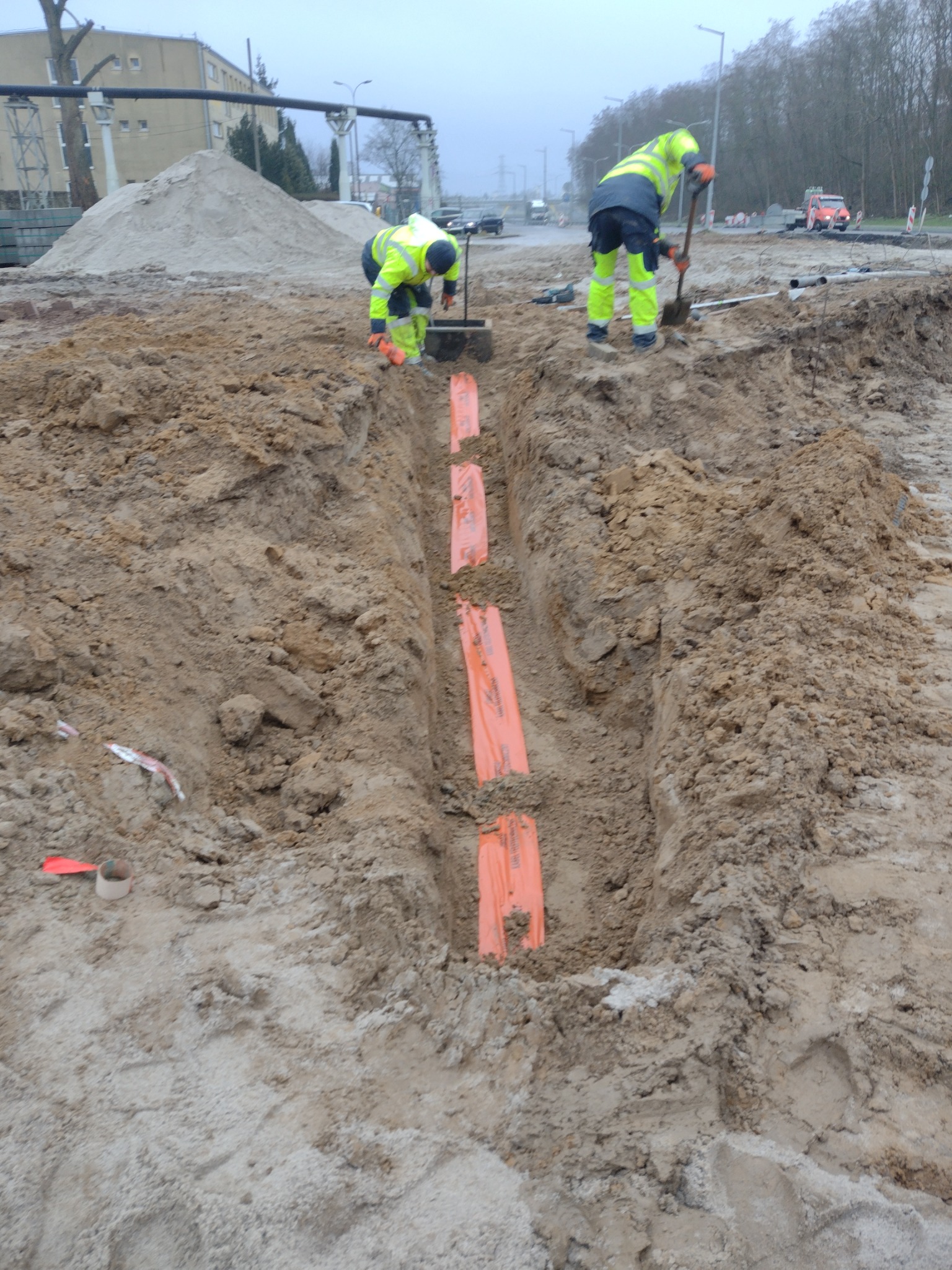 Dwóch pracowników w odblaskowych ubraniach układa pomarańczowe rury w wykopie ziemnym, w tle hałda piasku i ruch uliczny.