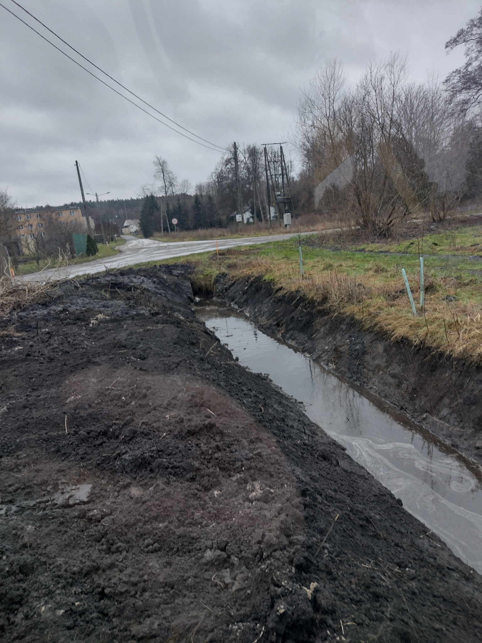 Wykopany rów melioracyjny wypełniony wodą, z widoczną świeżo usypaną ziemią po bokach, w tle droga i zabudowania w pochmurny dzień.