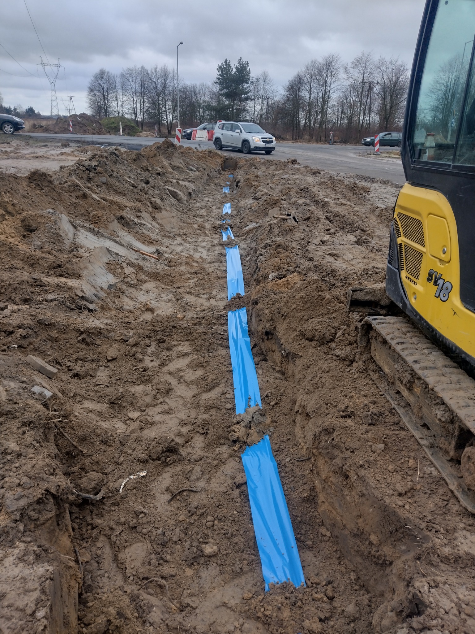 Wykopany rów z widocznymi niebieskimi rurami osłonowymi, częściowo zasypany ziemią, obok żółta koparka, w tle droga i drzewa pod pochmurnym niebem.