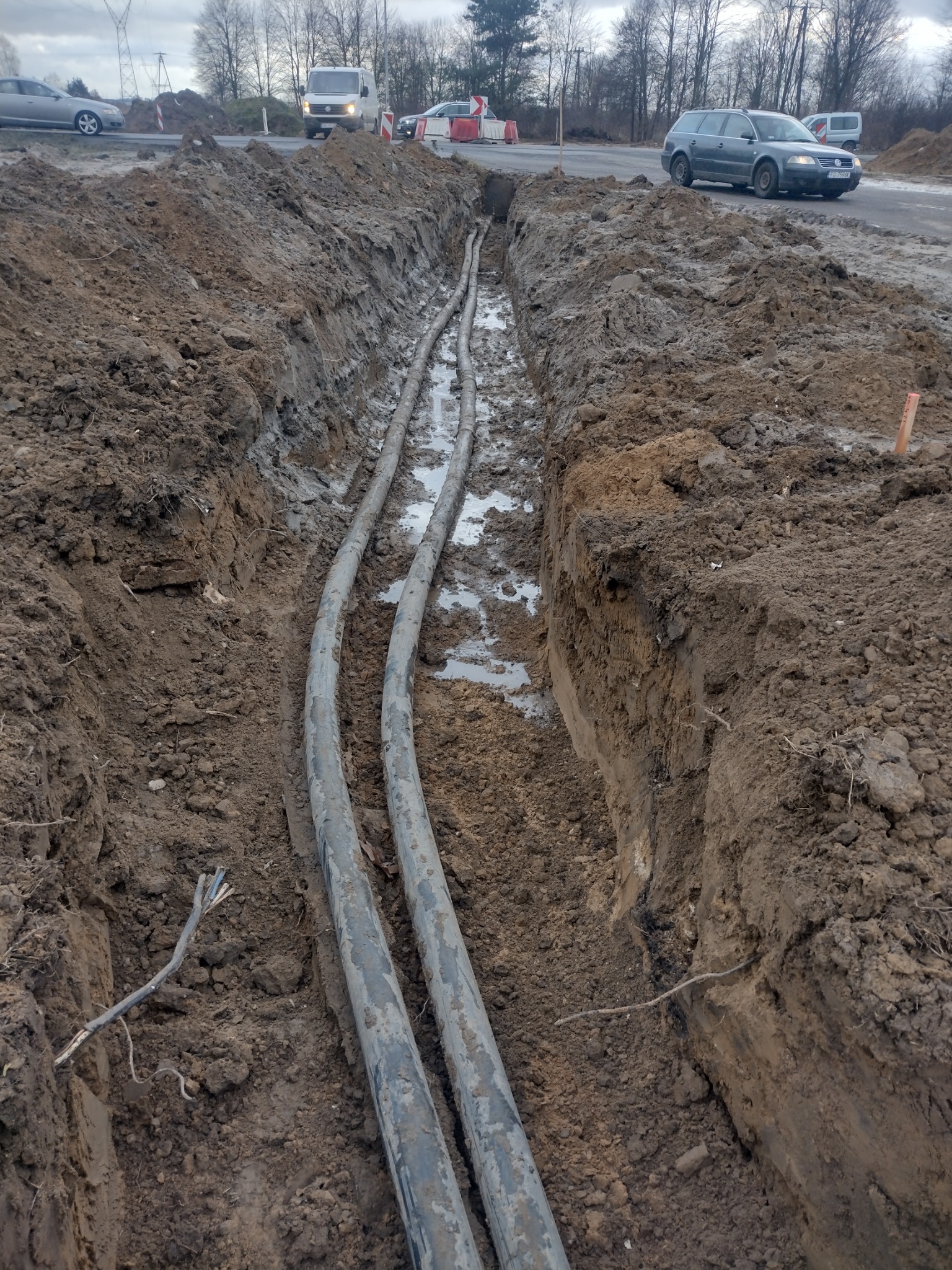 Dwa czarne kable ułożone w wykopie ziemnym, częściowo wypełnionym wodą, z widocznymi samochodami i drzewami w tle.