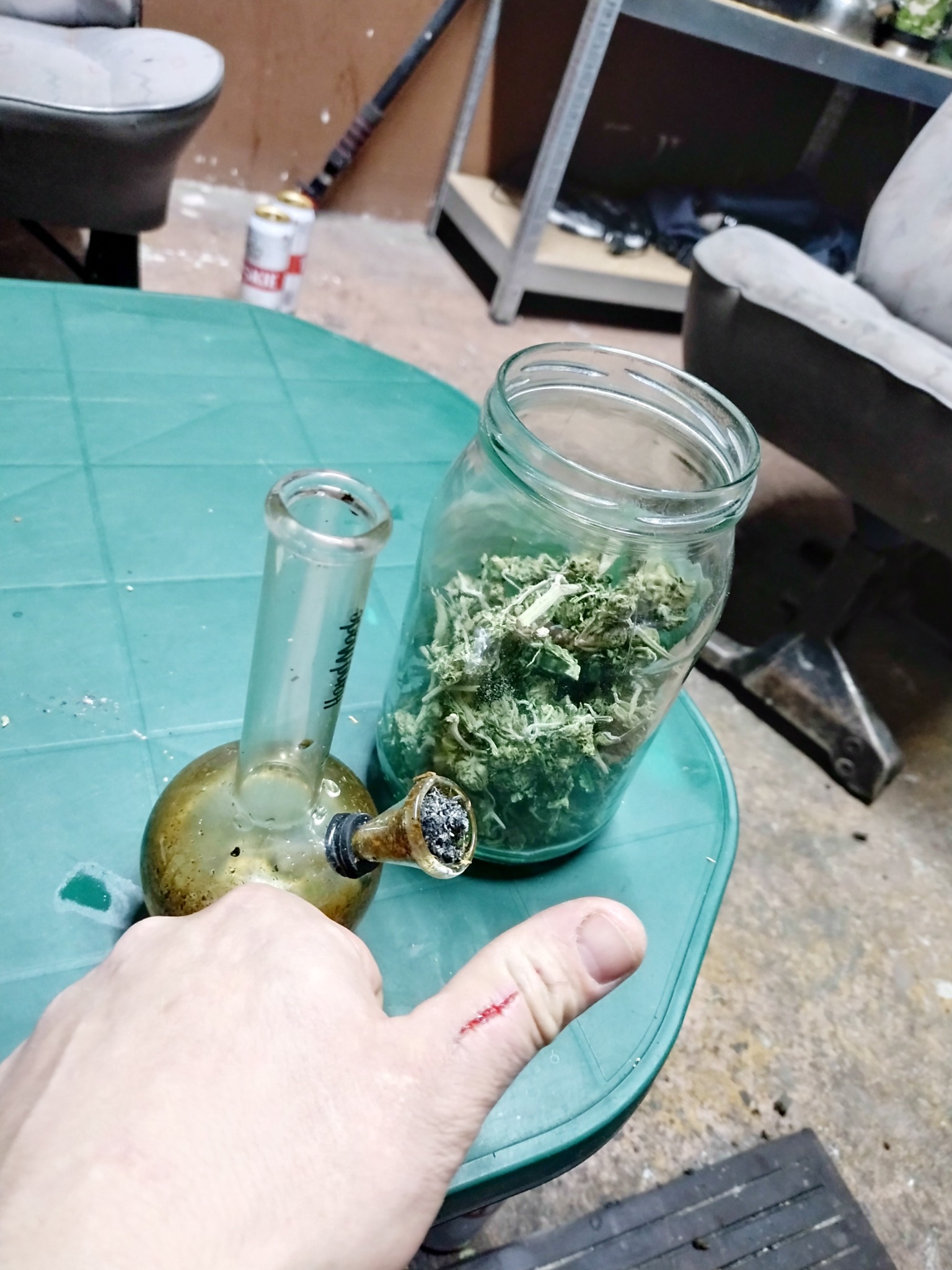 Brudny bongo i słoik z suszem na zielonym stole, widoczna ręka z zadrapaniem na kciuku, w tle puszki po piwie i regał.