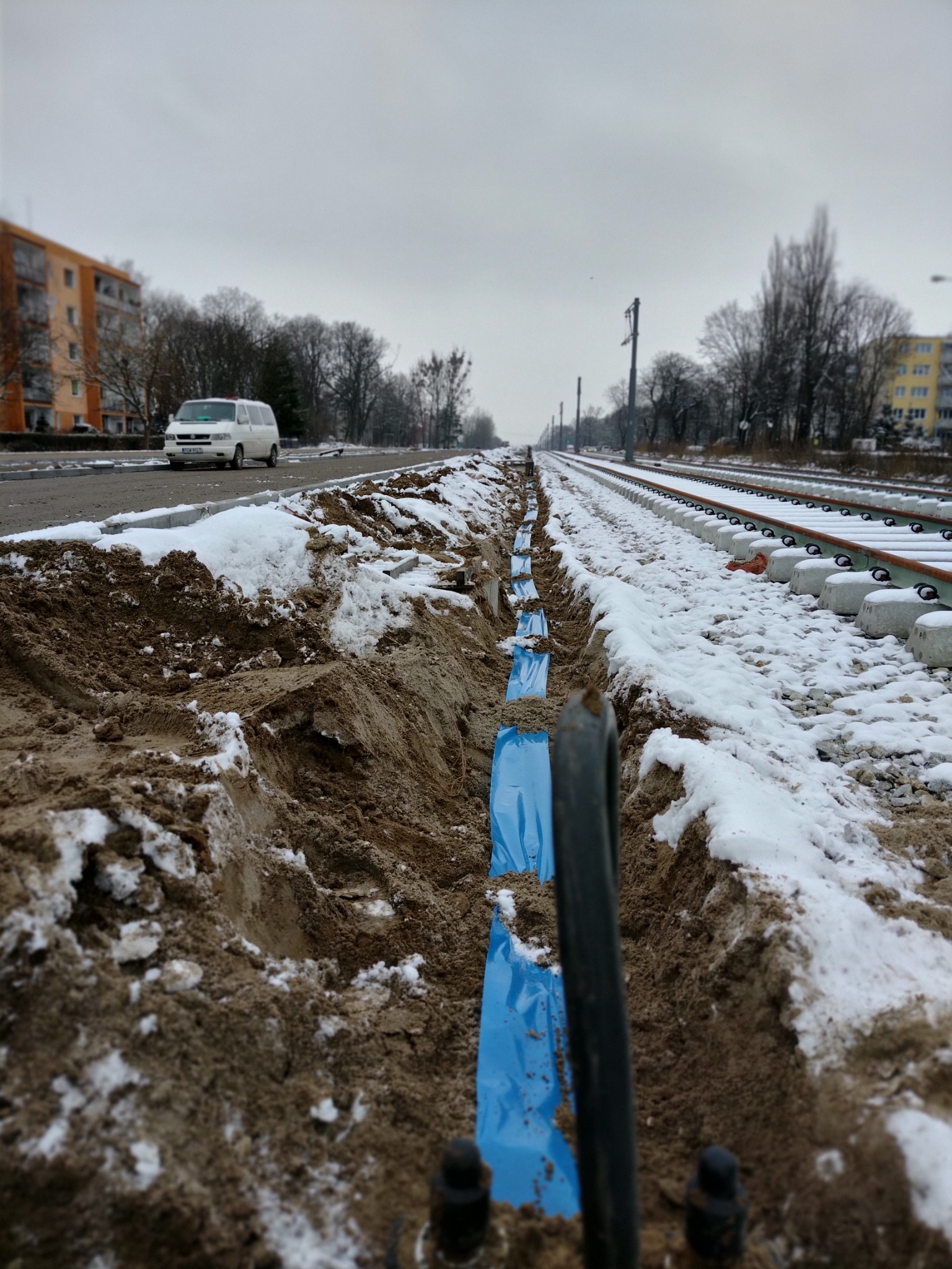 Wykopany rów w ziemi, częściowo zasypany śniegiem, zabezpieczony niebieską folią, biegnący wzdłuż torów kolejowych, w tle budynki mieszkalne i biały van.