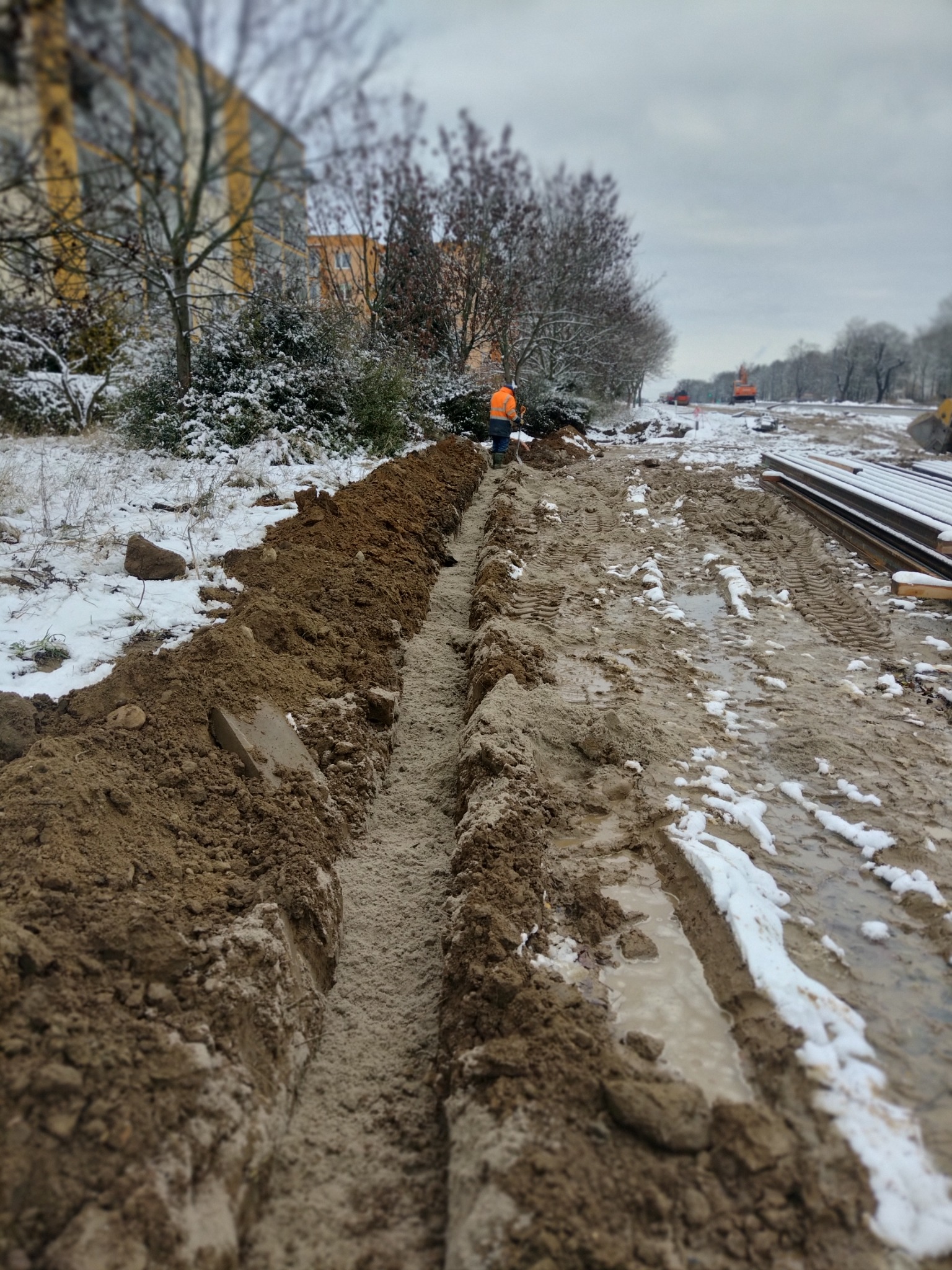 Długi, wąski wykop w ziemi pokrytej śniegiem, z pracownikiem w pomarańczowej kamizelce i ciężkim sprzętem w oddali.