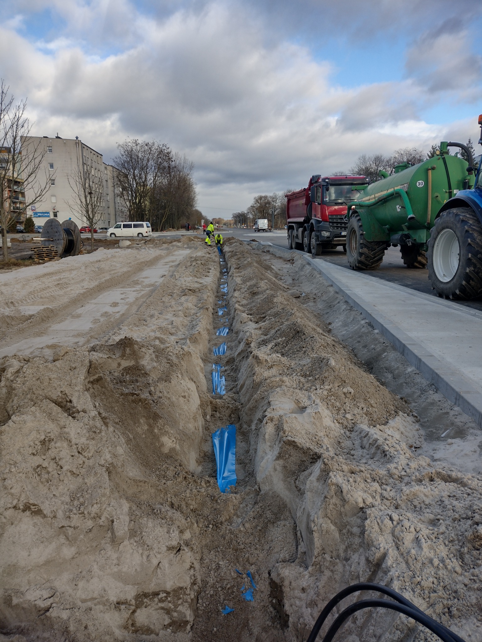 Wykopany rów w piasku z niebieskimi osłonami kabli, pracownicy w odblaskowych kamizelkach, ciężarówka i traktor w tle, miejski krajobraz.