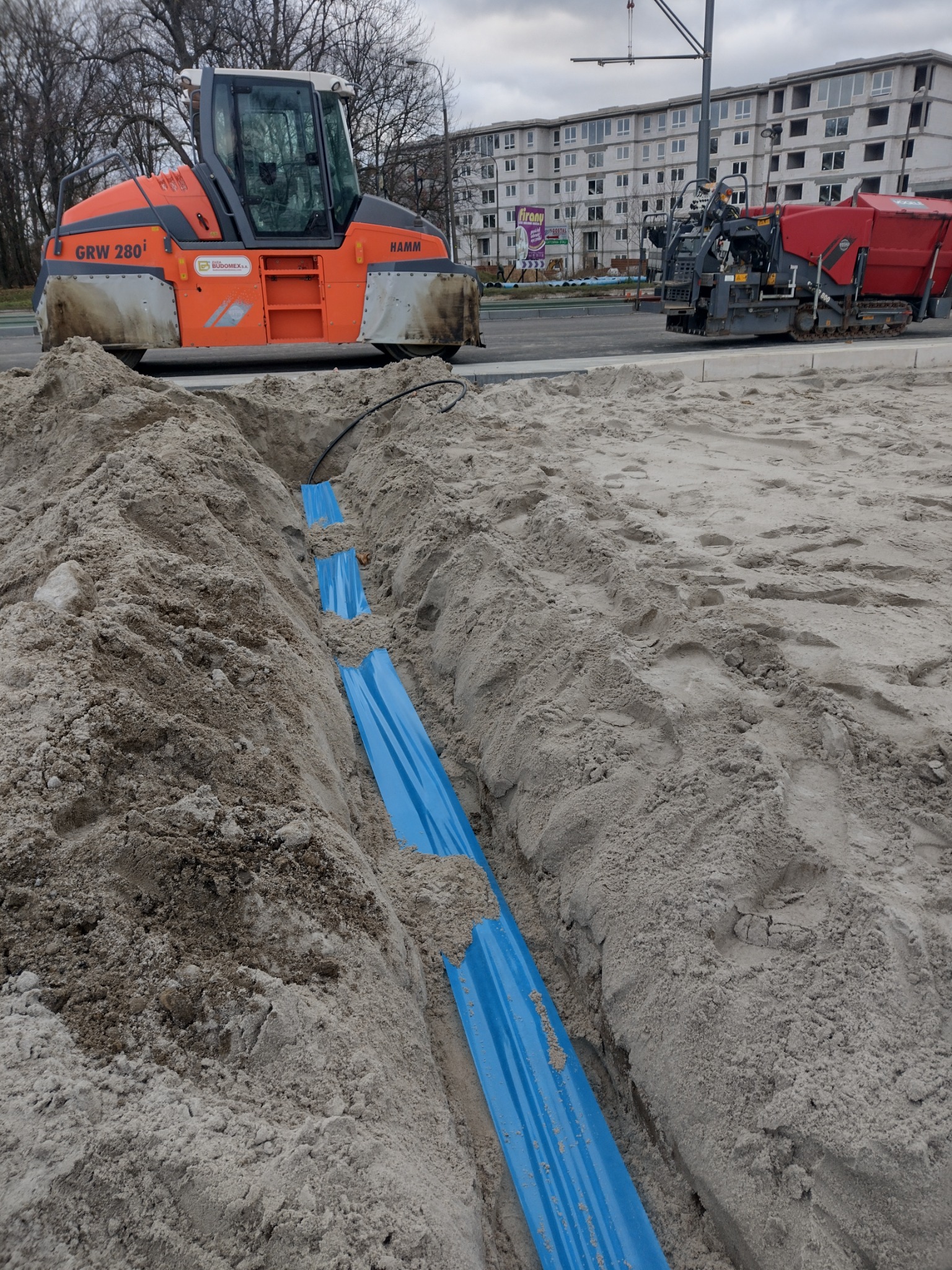 Wykop w piasku z ułożonymi niebieskimi rurami ochronnymi, w tle walec drogowy Hamm GRW 280i i rozściełacz asfaltu Vogele Super 800.