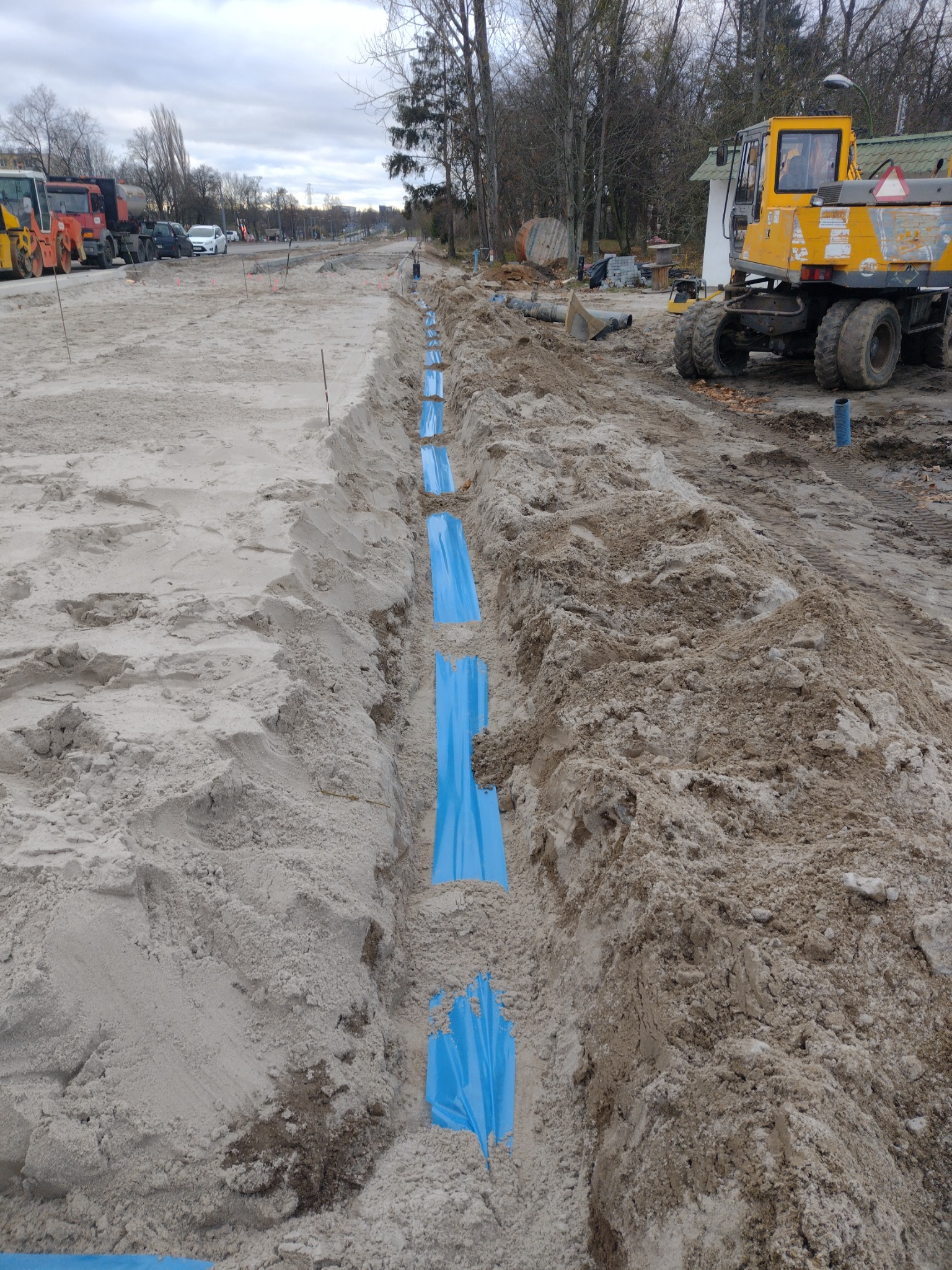 Wykopany rów z ułożonymi niebieskimi foliami ochronnymi, obok stoi żółta koparka, w tle inne maszyny budowlane i ulica.