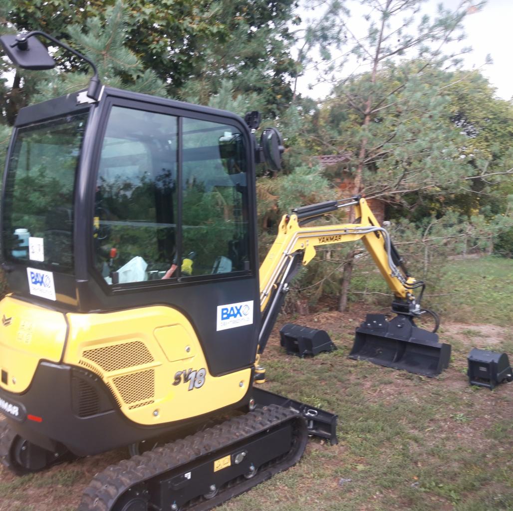 Żółto-czarna mini koparka Yanmar SV18 z łyżką na trawiastym terenie, obok leżą dwie czarne osłony. Widoczne drzewa w tle i logo BAX na maszynie.