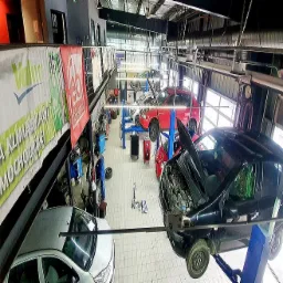 Warsztat samochodowy z podnośnikami, kilka aut w trakcie naprawy, otwarte maski, widok z góry, banery reklamowe na ścianach, narzędzia na posadzce.