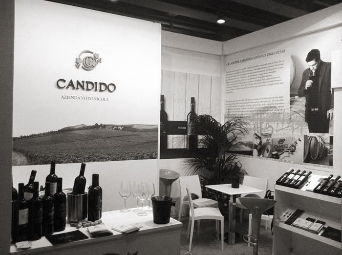 Czarno-białe zdjęcie stoiska firmy winiarskiej z butelkami wina, kieliszkami, stolikiem i grafikami przedstawiającymi winnice i degustację wina.