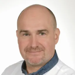 Portret mężczyzny z krótką brodą, w białej koszuli z dżinsowym kołnierzem, na jasnym tle.