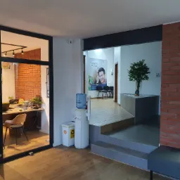 Wnętrze szkoły językowej z recepcją widoczną przez przeszklone drzwi, w tle plakat z napisem 'Metoda BLS uczy mówić' oraz drzewko bonsai w minimalistycznej donicy.