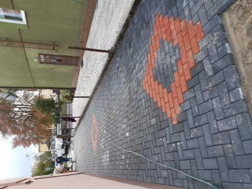 Nowo ułożona ścieżka z szarej kostki brukowej z wzorem rombu z czerwonej kostki, widoczna krawędź trawnika i fragment budynku w tle.