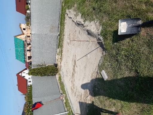 Przygotowanie podłoża pod nawierzchnię z kostki brukowej, widoczna warstwa piasku, szpadel wbity w trawę na skraju terenu, oraz budynki mieszkalne w tle.