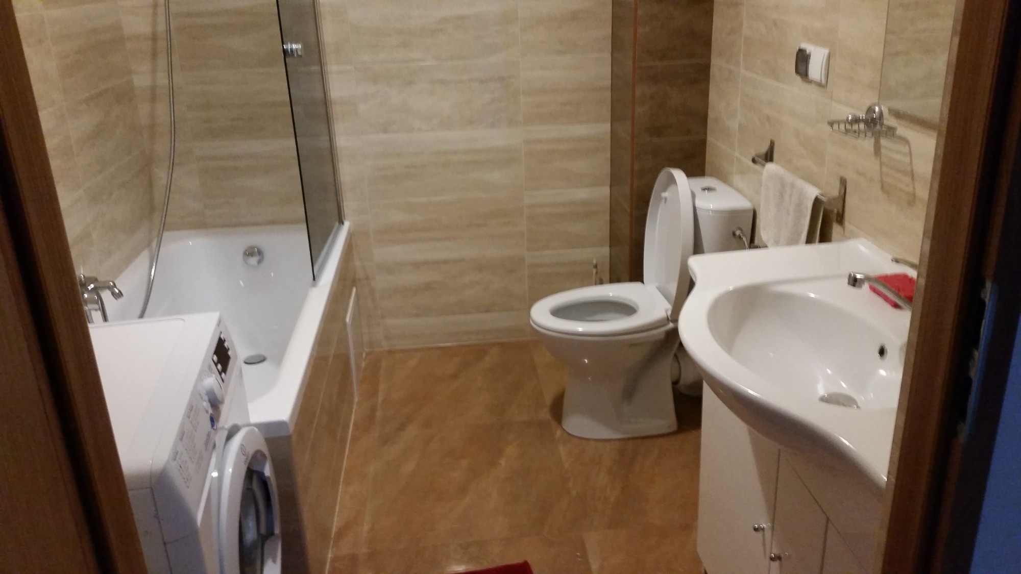 Wnętrze łazienki z beżowymi płytkami, obejmujące umywalkę z szafką, toaletę, wannę z parawanem oraz pralkę, całość utrzymana w ciepłej kolorystyce.