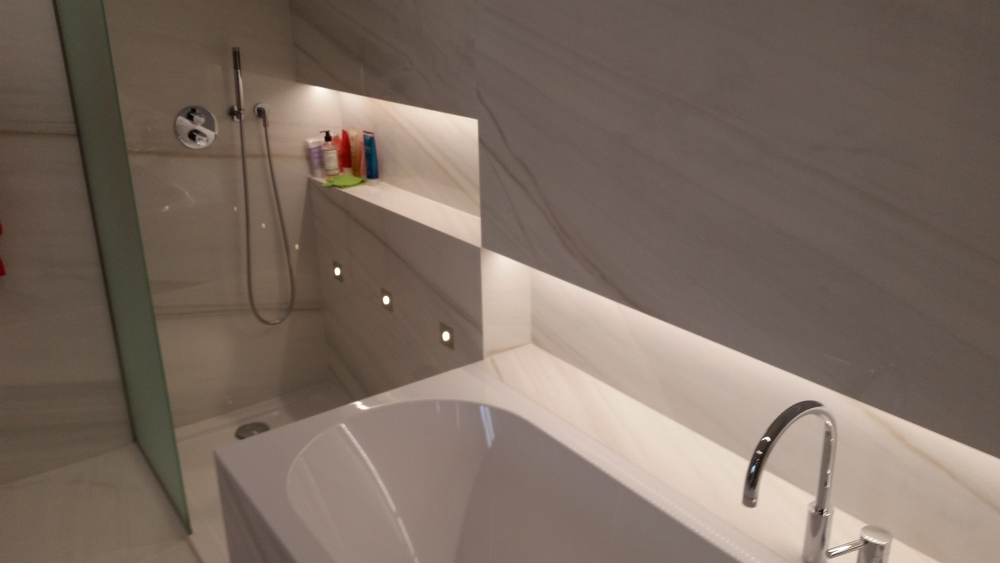 Nowoczesna łazienka z wanną, kabiną prysznicową, półkami wnękowymi z oświetleniem punktowym i marmurowymi płytkami.