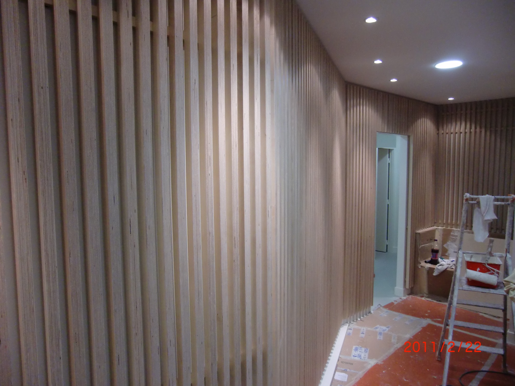 Wykończenie ścian w biurze pionowymi listwami z jasnego drewna, widoczny fragment podłogi zabezpieczonej kartonem i drabina malarska w tle.