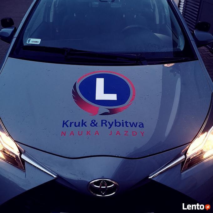 Szary samochód z naklejką z literą L i nazwą firmy 'Kruk & Rybitwa Nauka Jazdy' na masce, widok z przodu, lekko z góry.