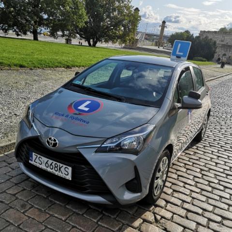 Szary samochód Toyota Yaris z oznakowaniem szkoły jazdy 'Kruk & Rybitwa' oraz niebieską literą 'L' na dachu, zaparkowany na brukowanej ulicy z widokiem na park i most w tle.