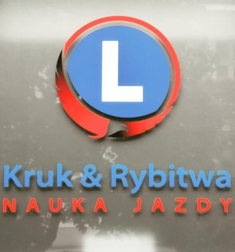 Szyld z logo firmy 'Kruk & Rybitwa' oferującej naukę jazdy, z charakterystycznym symbolem 'L' w niebiesko-czerwonym okręgu.