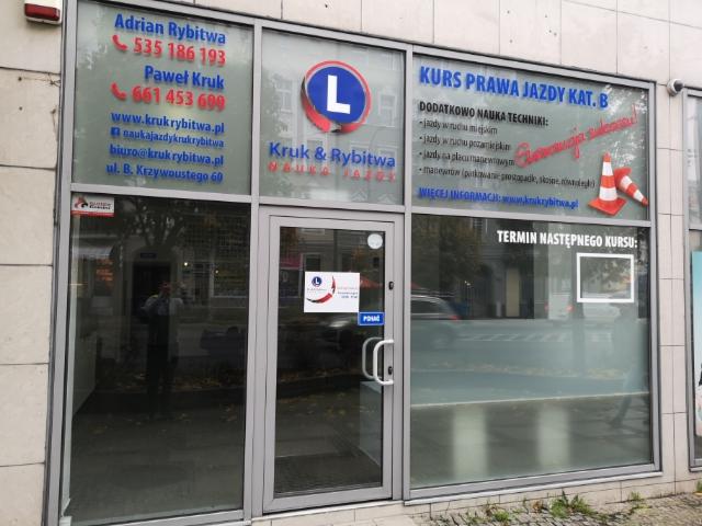 Wejście do firmy Kruk & Rybitwa z naklejonymi informacjami o kursach prawa jazdy kategorii B, numerami telefonów kontaktowych i adresem strony internetowej.