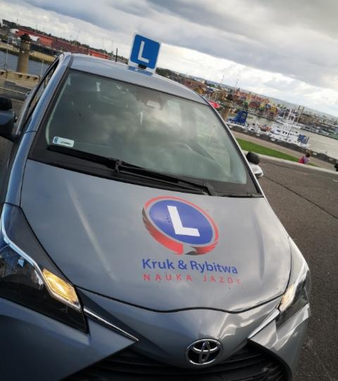 Szary samochód oznaczony literą 'L' na dachu i masce, z nazwą 'Kruk & Rybitwa Nauka Jazdy', w tle widok na port.