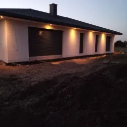 Nowoczesny dom jednorodzinny z płaskim dachem, oświetlony punktowymi lampami elewacyjnymi, widoczne niedokończone prace ziemne wokół budynku.