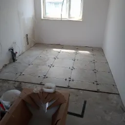 Układanie beżowych płytek podłogowych w jasnym pomieszczeniu, widoczne krzyżyki dystansowe, przygotowane narzędzia i materiały, ściany częściowo wykończone.