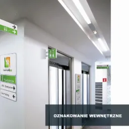 Wnętrze biurowca z systemem oznakowania: podwieszane tablice kierunkowe, tabliczki informacyjne na ścianie, wskazujące kierunki do biur i toalet.