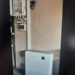 Business Consulting Solutions Krzysztof Strug - Biały system magazynowania energii SIGENENERGY z falownikiem, zainstalowany obok otwartej rozdzielnicy elektrycznej na beżowej ścianie.