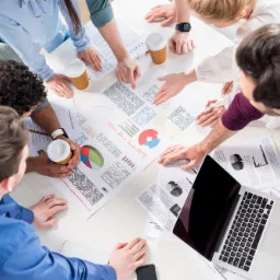 Zespół osób pochylony nad stołem z wydrukami wykresów i laptopem, analizujący dane podczas spotkania, widok z góry.
