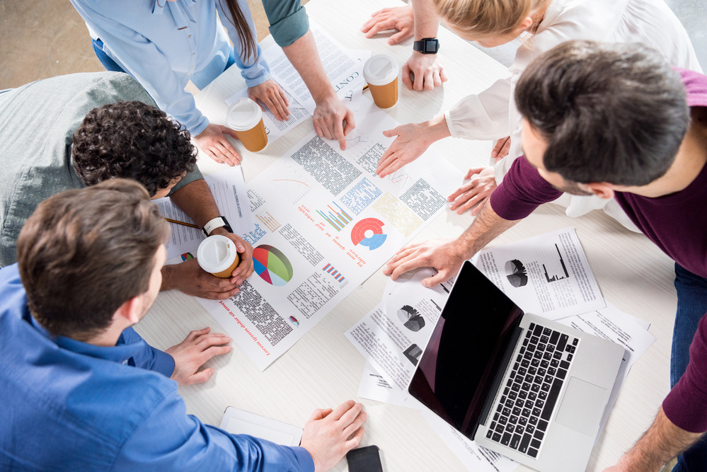 Zespół osób pochylony nad stołem z wydrukami wykresów i laptopem, analizujący dane podczas spotkania, widok z góry.