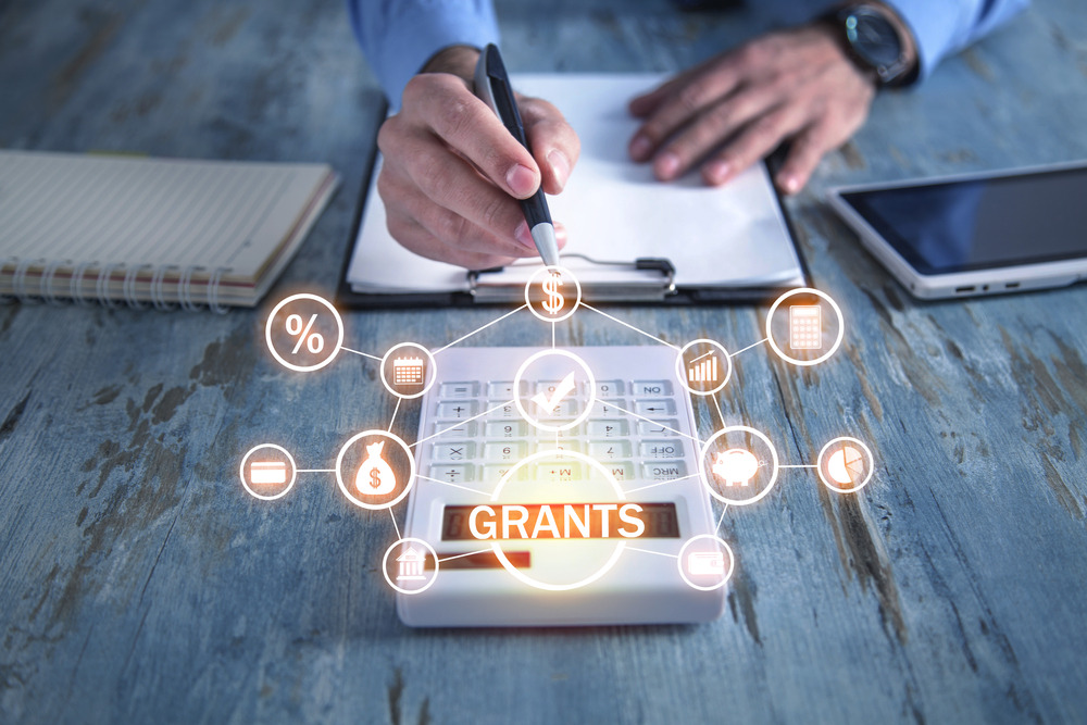 Kalkulator z napisem GRANTS otoczony ikonami finansowymi na drewnianym biurku, dłoń z długopisem nad dokumentem, tablet i notatnik w tle.