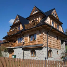 Tradycyjny, drewniany dom góralski z kamienną podmurówką, czarnym dachem i balkonami, otoczony drewnianym płotem, w słoneczny dzień.