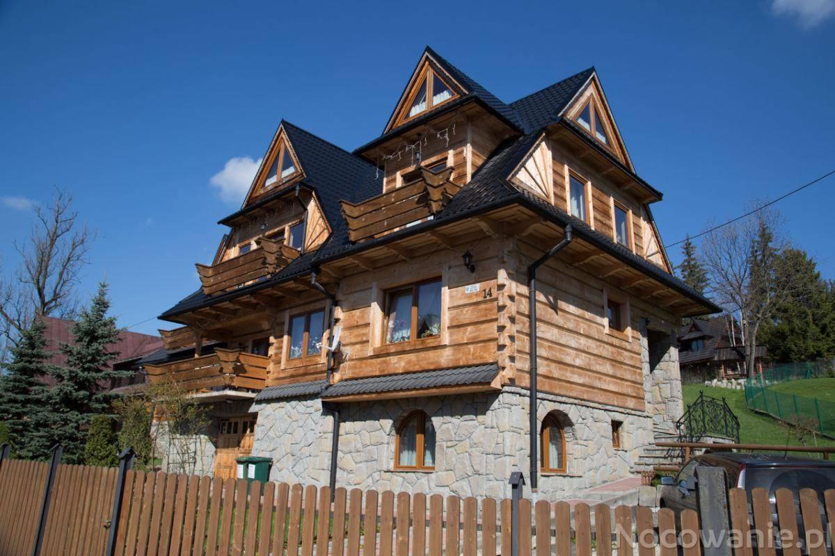 Tradycyjny, drewniany dom góralski z kamienną podmurówką, czarnym dachem i balkonami, otoczony drewnianym płotem, w słoneczny dzień.