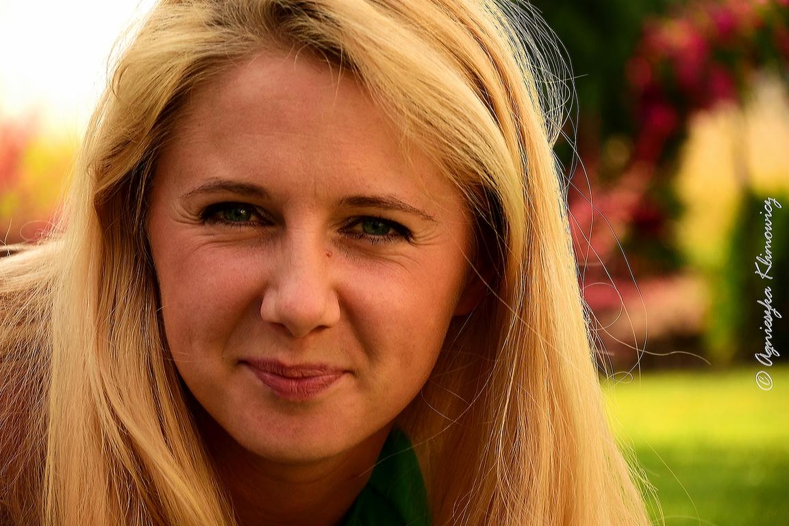 Portret kobiety z długimi blond włosami, zielonymi oczami i delikatnym uśmiechem, sfotografowana na tle rozmytej zieleni i kwiatów.