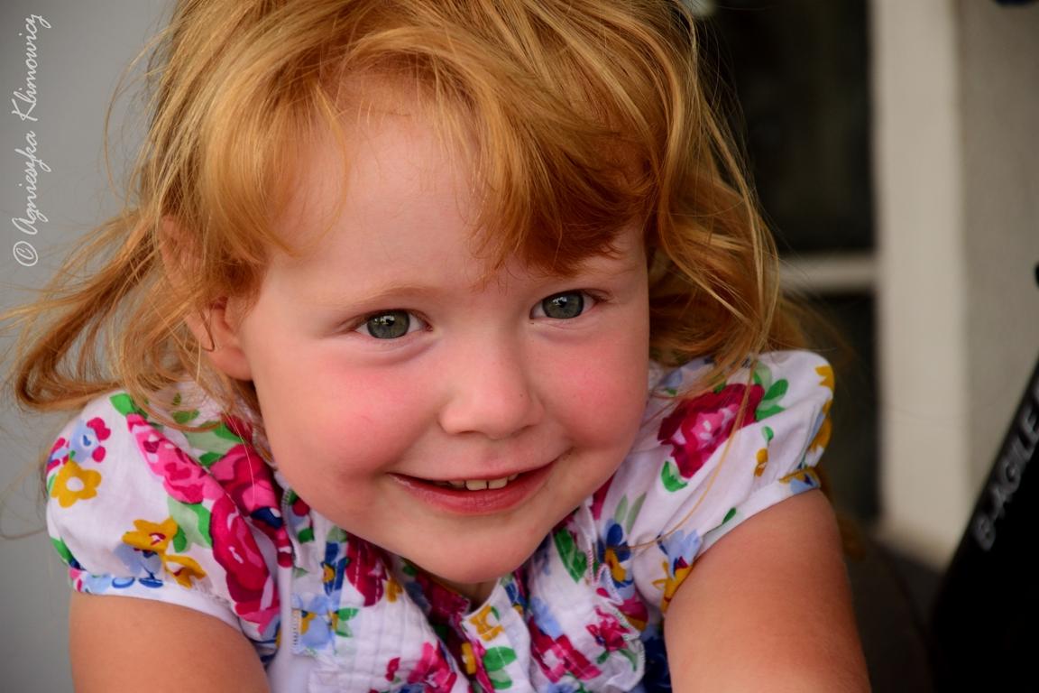 Portret uśmiechniętej, rudowłosej dziewczynki w kwiecistej bluzce, z delikatnym rumieńcem na policzkach i zielonymi oczami.