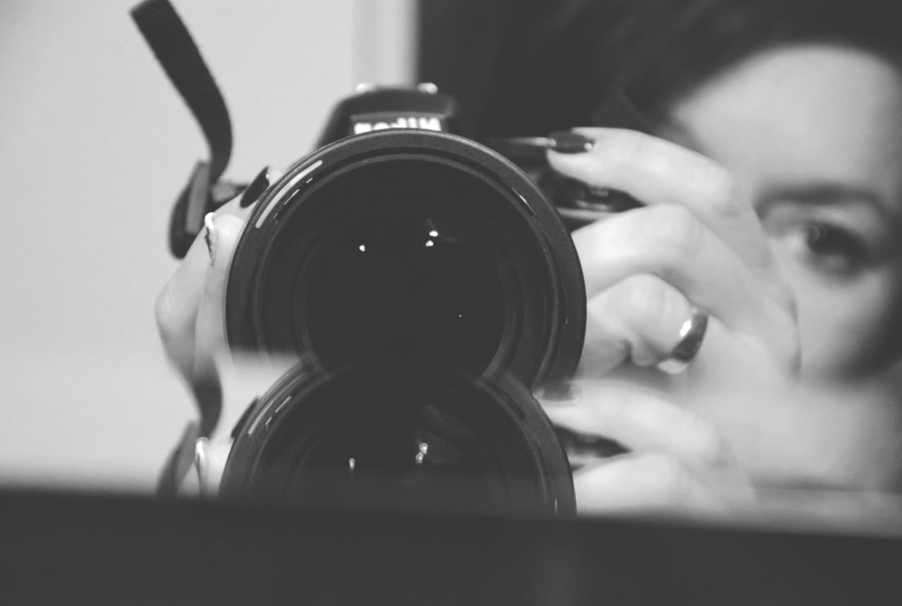 Czarno-białe ujęcie z bliska obiektywu aparatu, za którym widać kobiece dłonie z ciemnym manicure i pierścionkiem oraz fragment twarzy z intensywnym spojrzeniem.