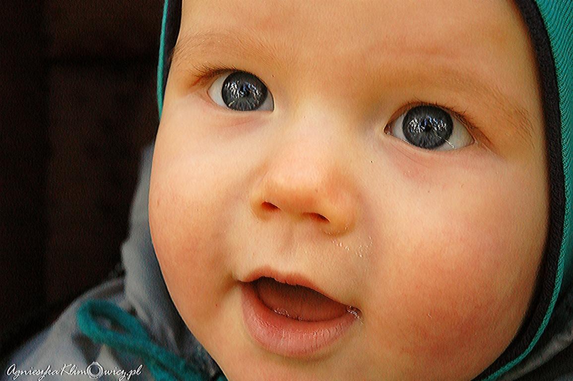 Zbliżenie na twarz uśmiechniętego niemowlęcia w czapce, z dużymi, ciemnymi oczami i delikatną skórą.