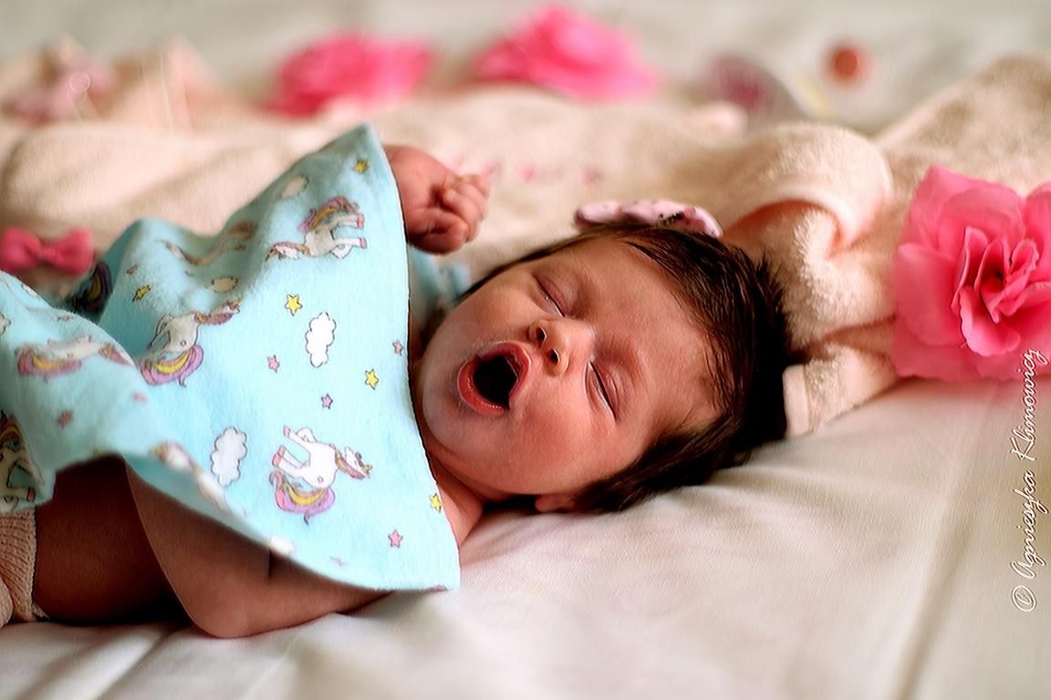 Śpiące niemowlę z otwartymi ustami, przykryte błękitnym kocykiem w jednorożce, obok różowa róża i dekoracyjne kwiatki.