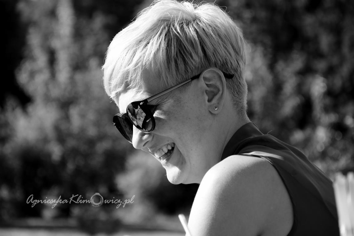 Portret uśmiechniętej kobiety w okularach przeciwsłonecznych w kształcie serc, z krótkimi włosami, wykonany w czerni i bieli na rozmytym tle zieleni.