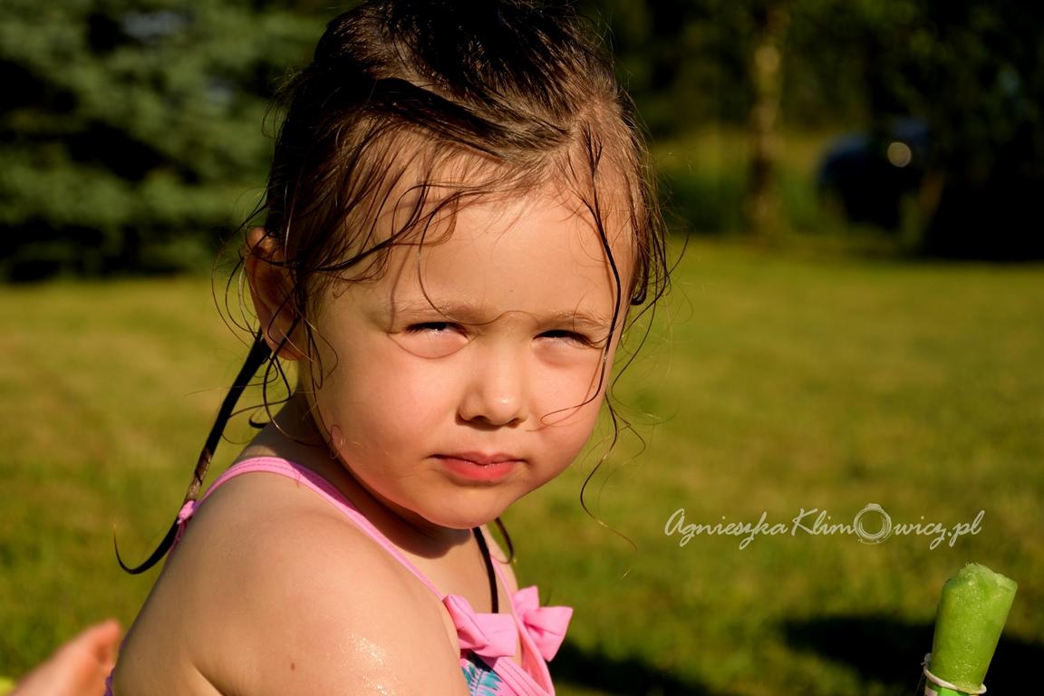 Portret dziewczynki z mokrymi włosami w letnim ubraniu na tle zielonego trawnika, z widocznym znakiem wodnym fotografa.