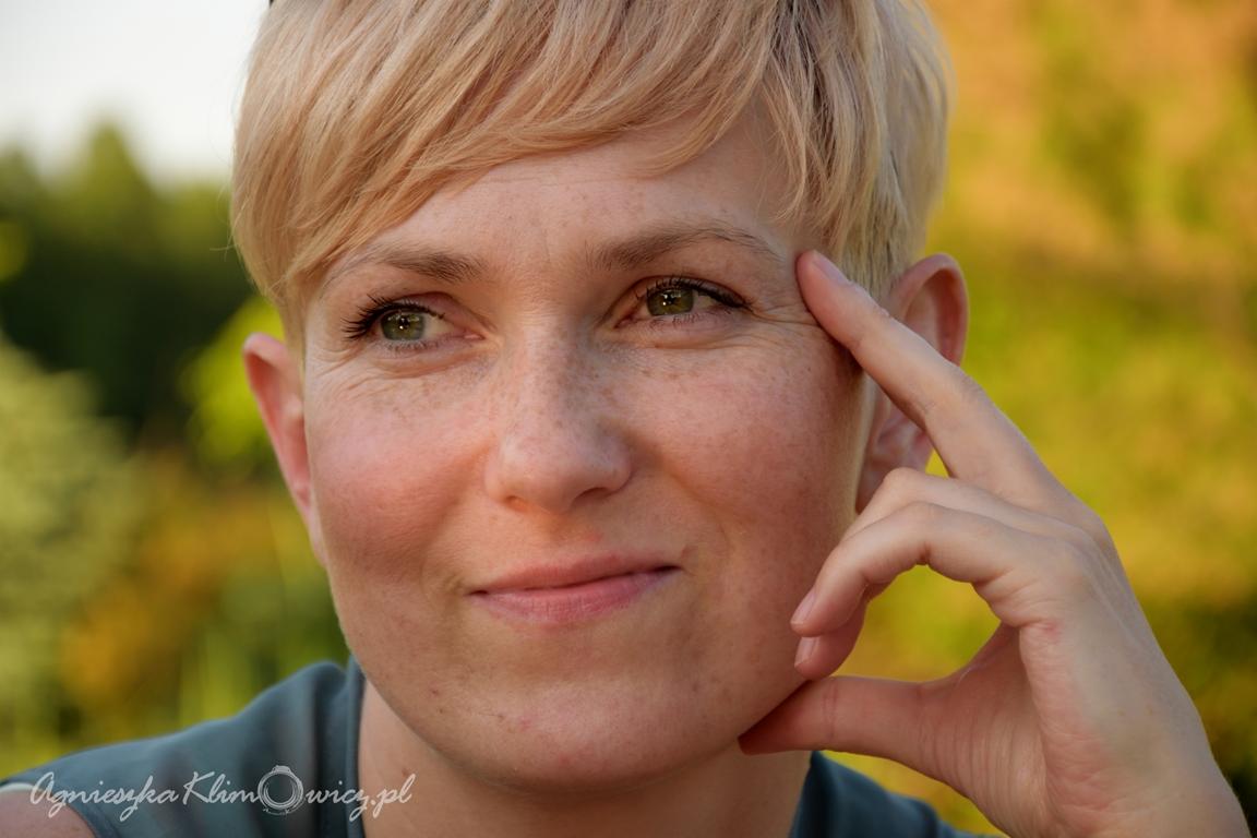 Portret uśmiechniętej kobiety z krótkimi blond włosami i piegami, opierającej palec o skroń na tle zieleni.