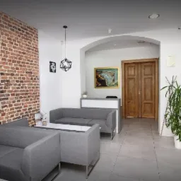 Wnętrze biura agencji nieruchomości z szarymi sofami, ceglaną ścianą z obrazem, recepcją w tle i logo firmy na ścianie.